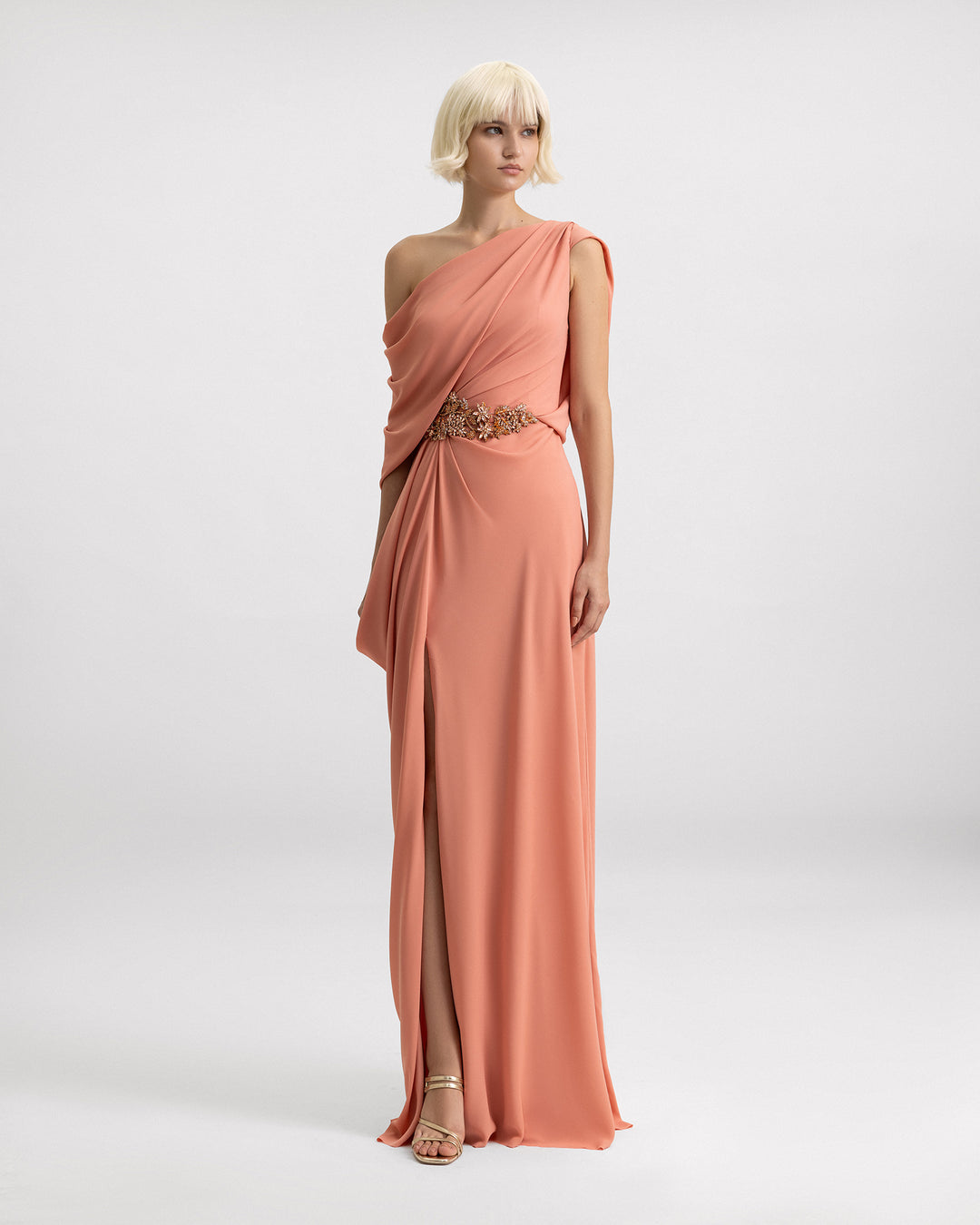 A one-shoulder draped evening dress in coral color with beadings on the waistline and a slit on the side.