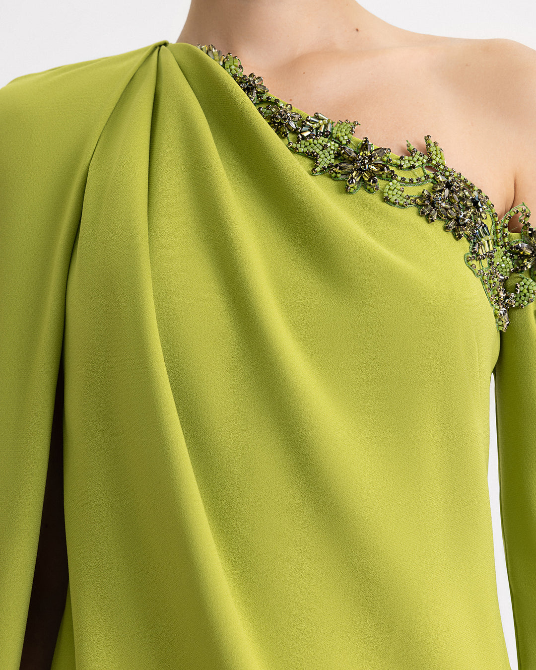 A close-up of a one-shoulder green evening dress with beadings on the neckline and a cape-like sleeve.