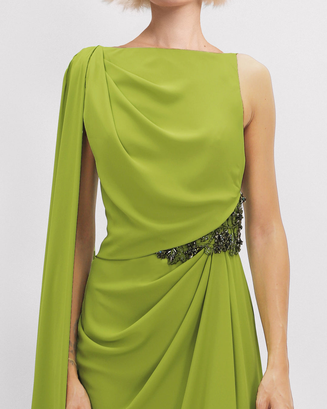 A close-up of an asymmetrical neckline evening dress with beadings on the waistline and a cape-like sleeve.