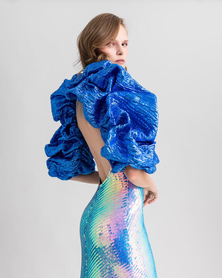 A close-up of a halter-neck slim-cut evening dress fully sequined in blue with a low open back, paired with a draped bolero.