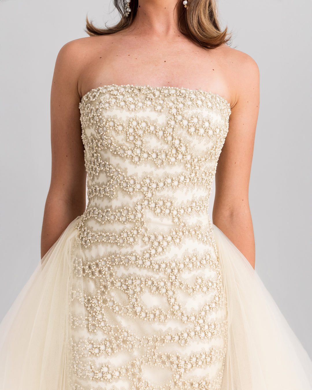 A close-up of a strapless, fully embroidered slim-cut evening dress in cream, with a flowy tulle tail.