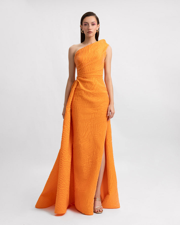 A one-shoulder evening dress in orange jacquard fabric with draping on the waistline and a slit on the side.