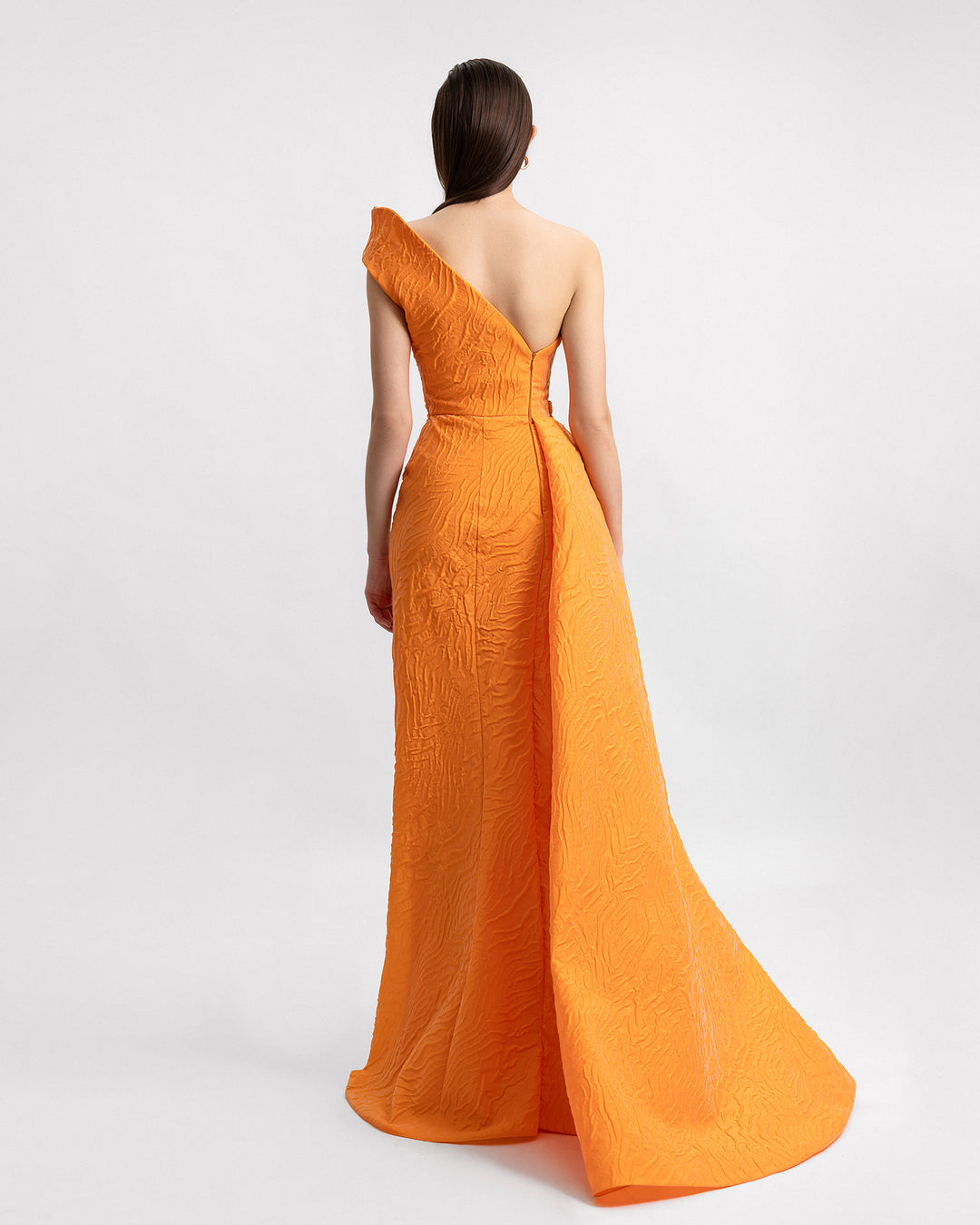 The back of a one-shoulder evening dress in orange jacquard fabric with draping on the waistline and an asymmetrical open back.