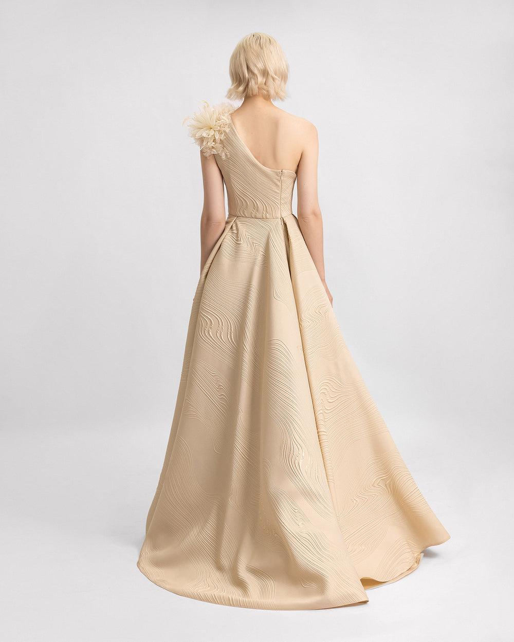 The back of a one-shoulder evening dress in beige jacquard with 3D flowers on the shoulder and a draped full skirt.