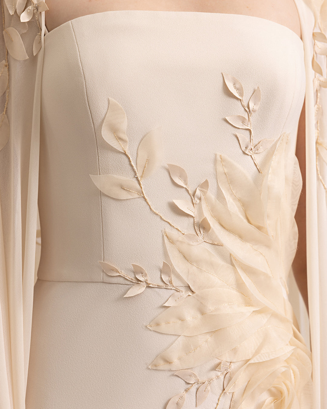 A close-up of a slim-cut champagne evening dress with floral details, paired with a long cape with embroidered organza flowers.