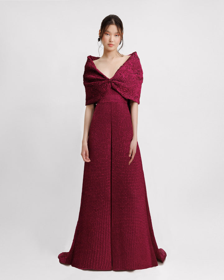 A dark fuchsia strapless evening dress with a plunging neckline and draping on the waistline, paired with a detachable bow-like cape.