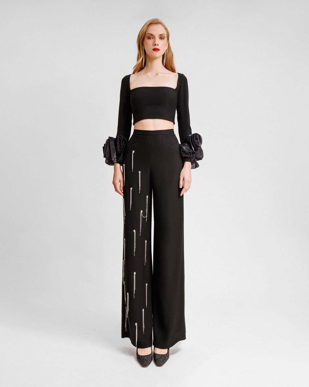 An evening wear set featuring a squared-neckline black top with draped floral cuffs, paired with a straight-cut pants with beading fringes on a single leg.