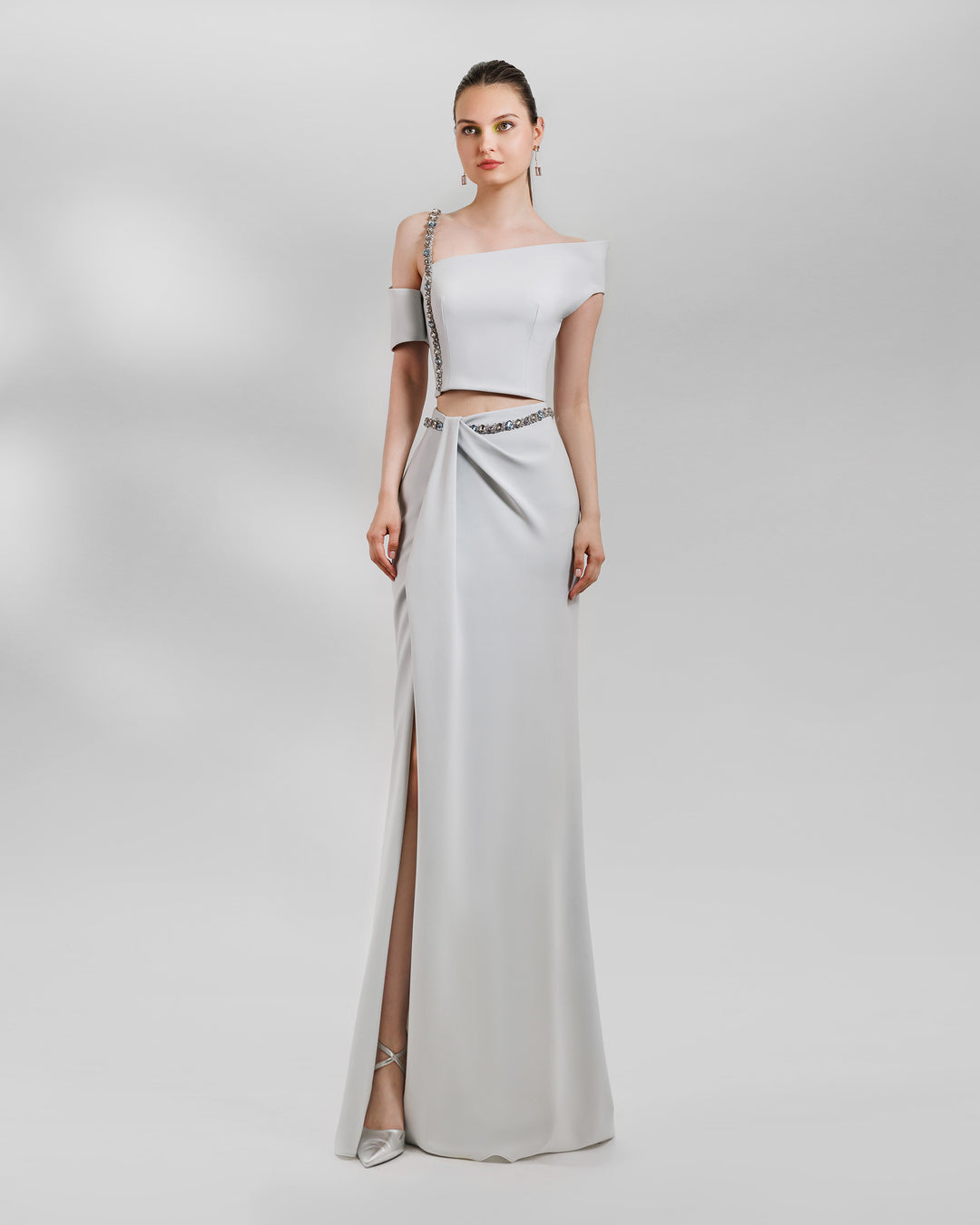 An evening wear grey set featuring an asymmetrical neckline top with a beaded strap paired with a draped skirt featuring a slit on the side and a detachable belt.