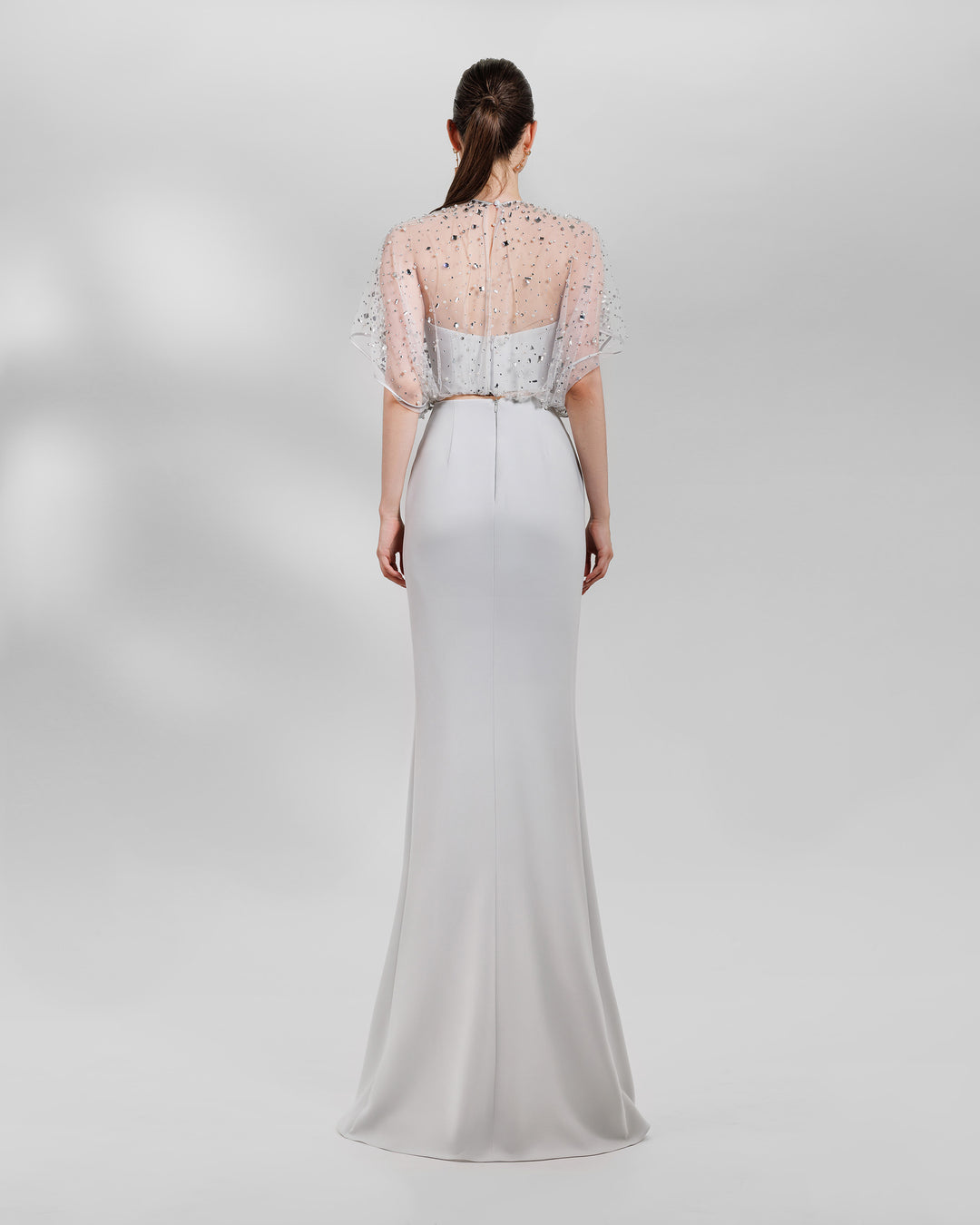 The back of an evening wear grey set featuring a fully beaded top with a crepe bow paired with a draped skirt featuring a slit on the side.