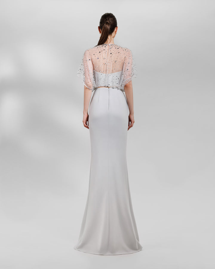 The back of an evening wear grey set featuring a fully beaded top with a crepe bow paired with a draped skirt featuring a slit on the side.