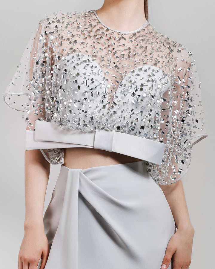 A close-up of an evening wear grey set featuring a fully beaded top with a crepe bow paired with a draped skirt featuring a slit on the side.