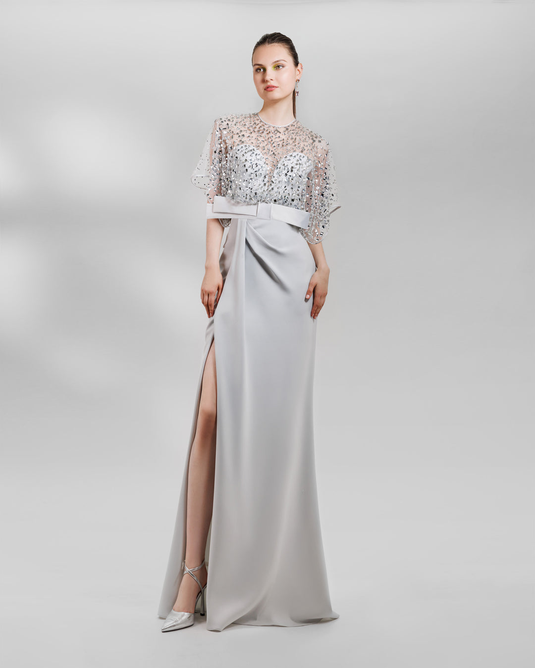 A long grey evening dress featuring a fully beaded top with a crepe bow paired with a draped skirt featuring a slit on the side.