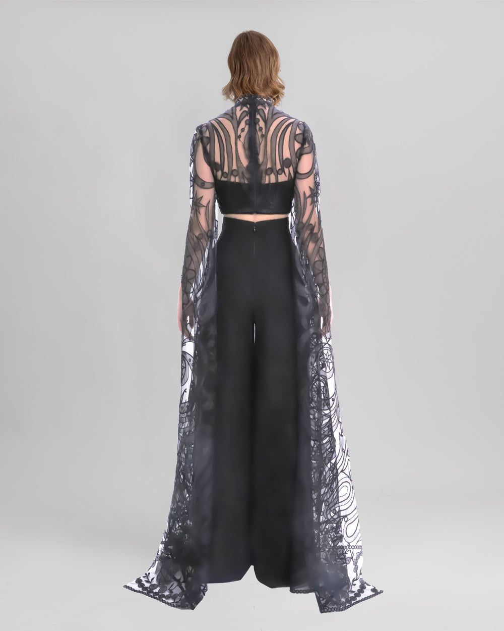 The back of an evening wear set featuring a black cropped lace top with floor-length sleeves, paired with a black pants featuring slits at the fronts.