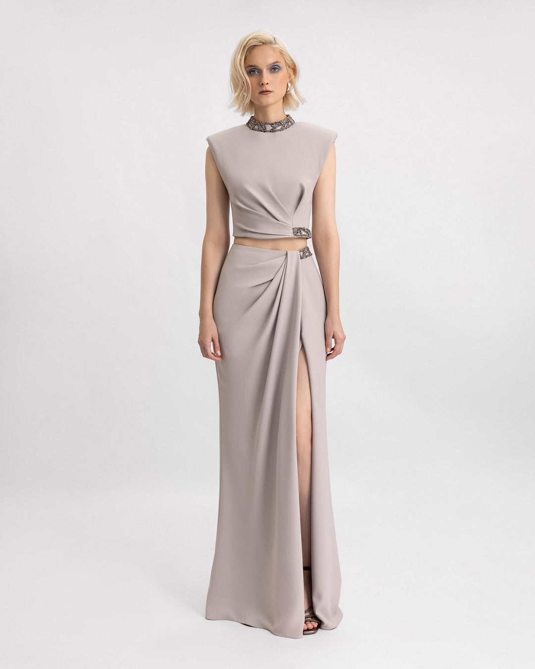 A grey evening wear set featuring beaded high-collar top with structured shoulders paired with a draped skirt with a slit on the side.