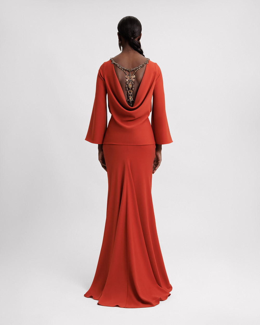 The back of an orange evening wear set featuring an cowl open back with a beaded bow design, paired on a long slim cut skirt.