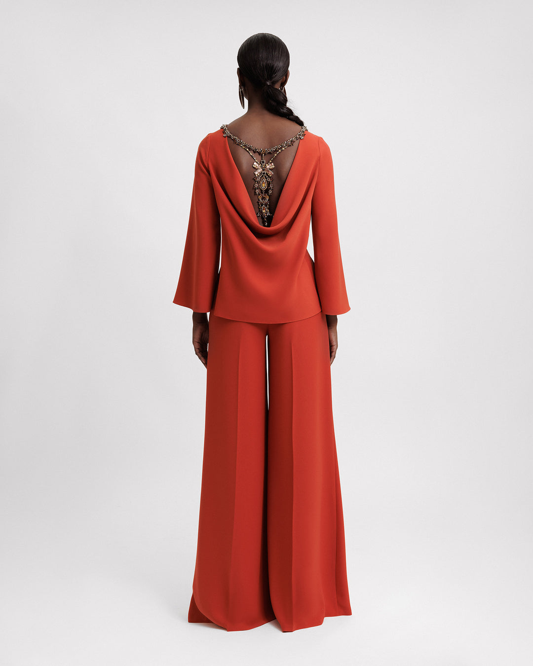 The back of an evening wear set featuring a long sleeve top with a beaded boat neckline and a beaded cowl open back, paired with a flared pant.