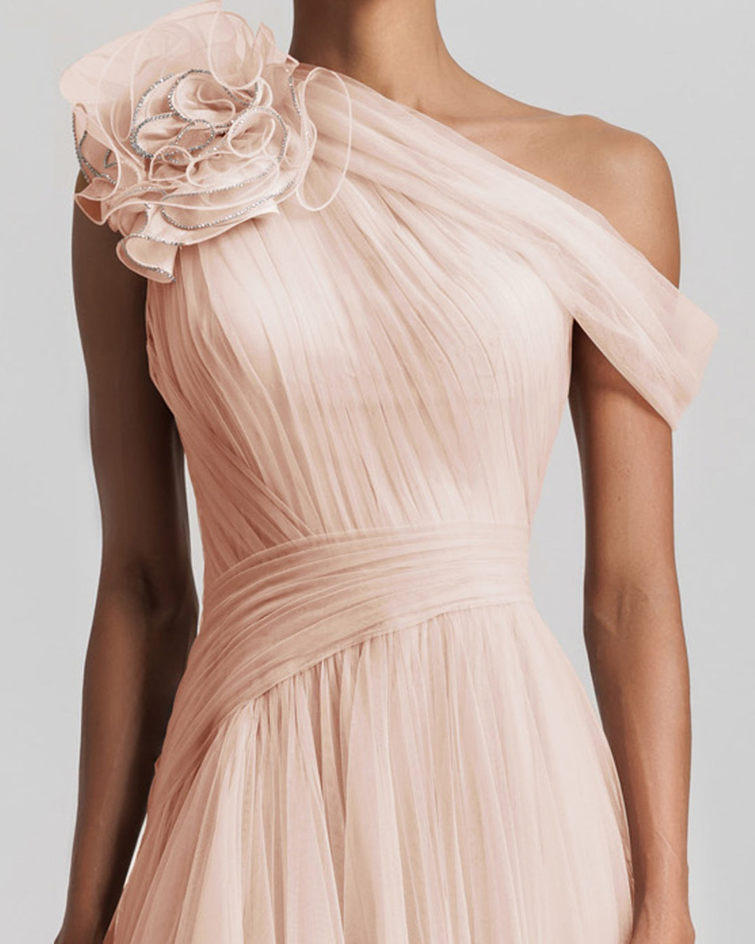 A close-up of an asymmetrical neckline full tulle long dress in blush color, with a flower design on the shoulder, ruffled side slit and draping design on the bodice.