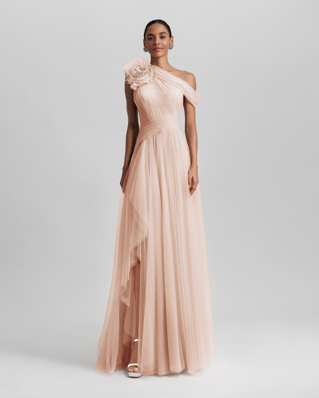 An asymmetrical neckline full tulle long dress in blush color, with a flower design on the shoulder, ruffled side slit and draping design on the bodice.