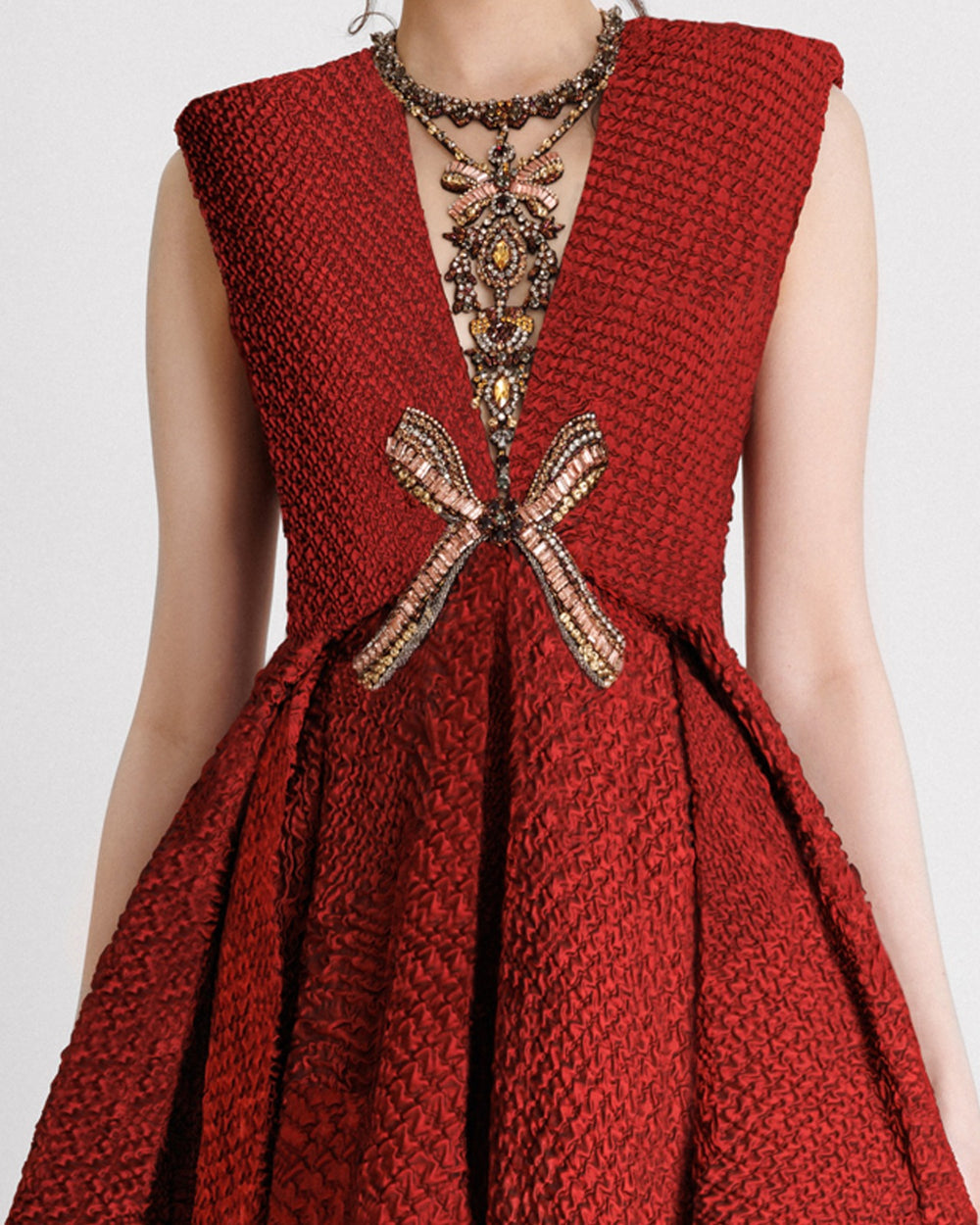 A close-up of an embellished V-neckline evening dress with structured shoulders and a draped flared skirt in red crinkled jacquard fabric.