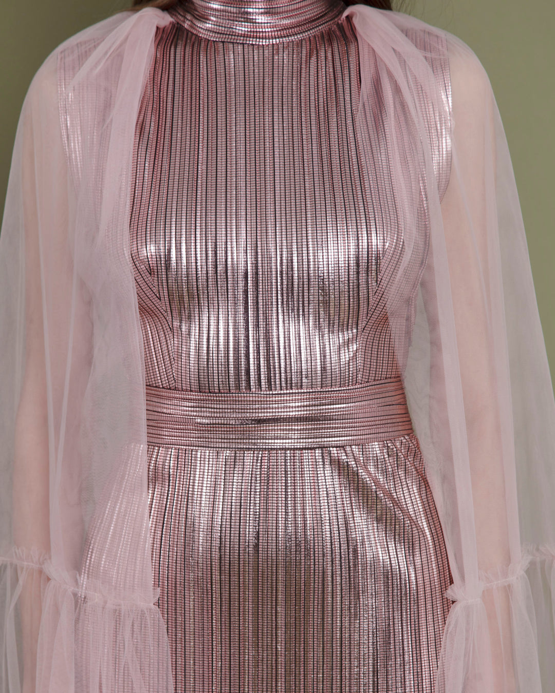 A close-up of a slim cut metallic pink evening dress in pleated fabric paired with a tulle ruffled cape.