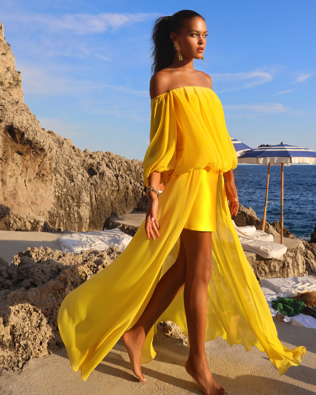 A model wearing a yellow off-shoulders, loose cut chiffon evening dress with an asymmetrical hemline at a capri beach.