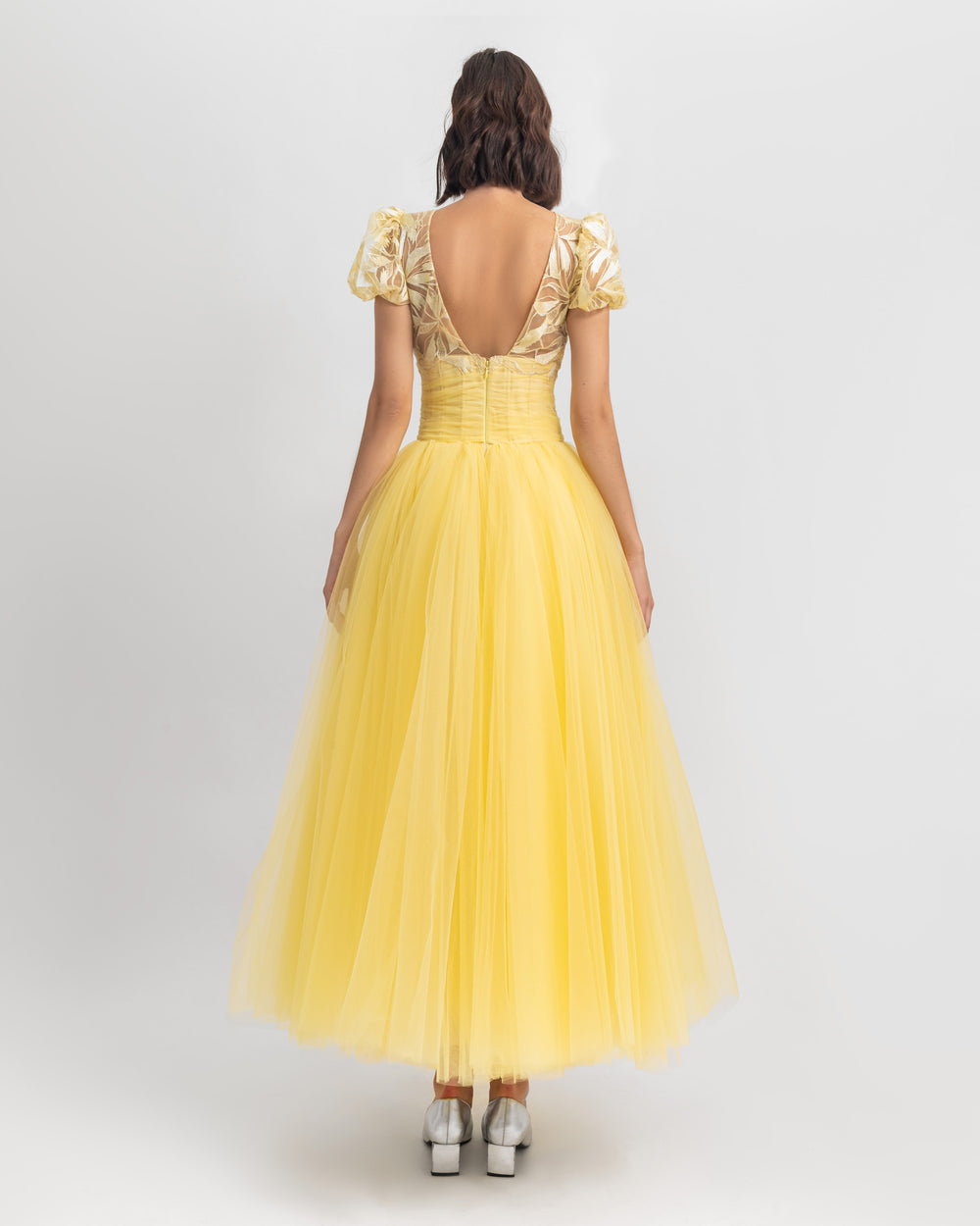 The back of a puffed sleeves intricated embroidery yellow evening dress with an open back and a wide cut tulle midi skirt.