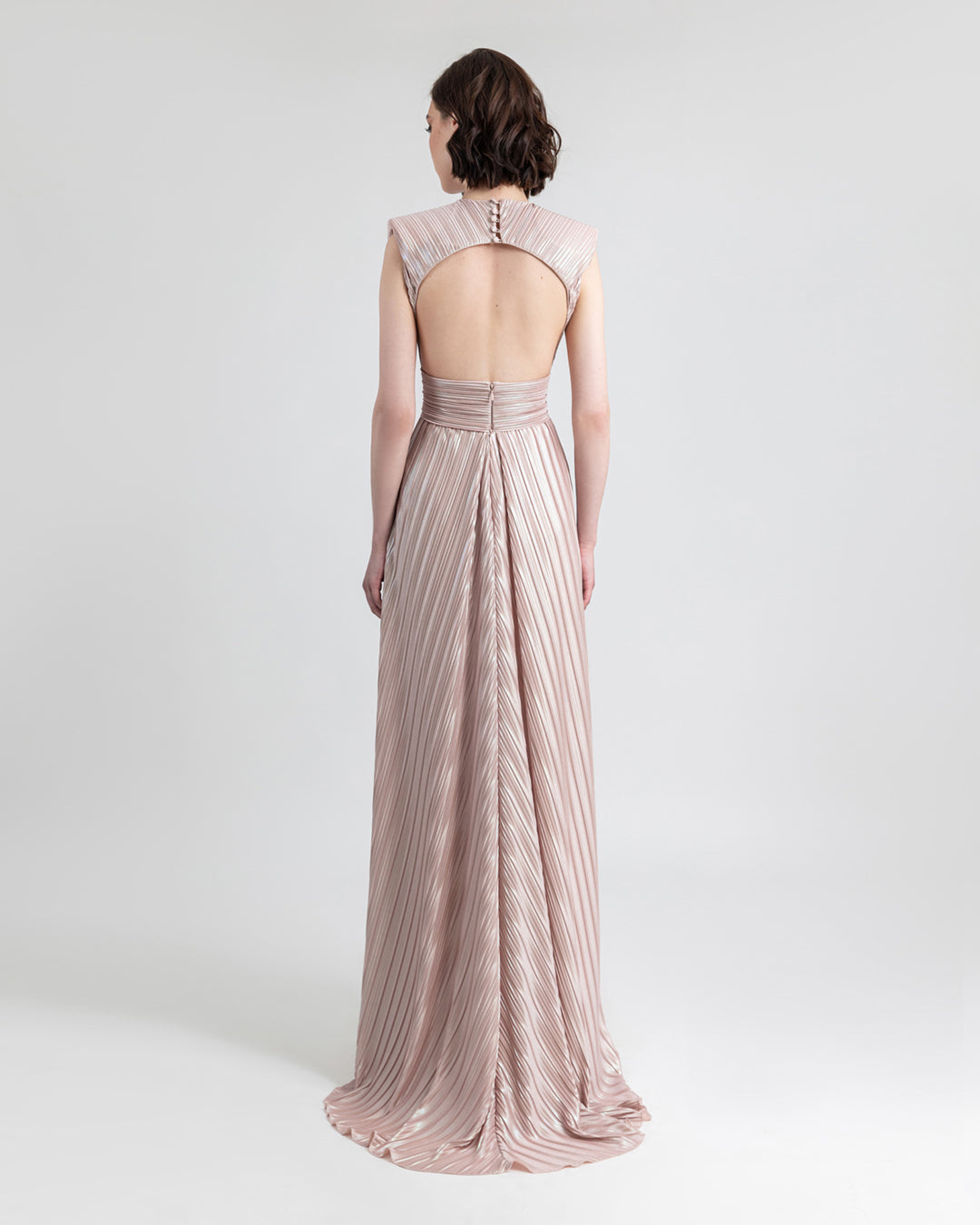 The back of a straight cut blush evening dress with ruffles details and open slit on the side in a pleated jersey fabric featuring an open back.