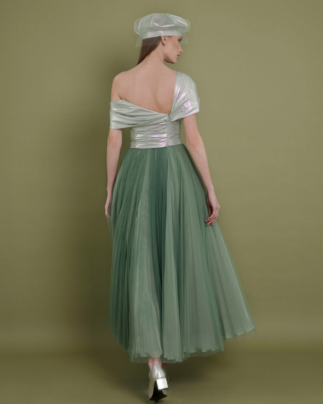 The back of a wide cut midi evening dress featuring an open back with asymmetrical neckline with metallic latex bodice and a pleated green tulle skirt.