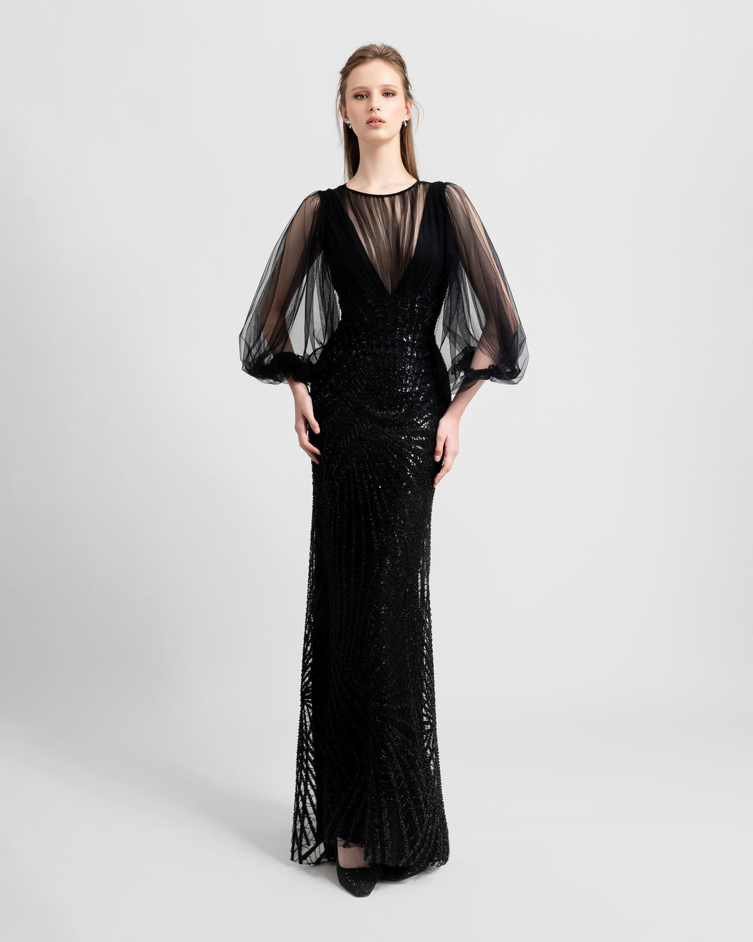 A deep V-neckline fitted black evening dress overlayed with a long sleeved draped tulle top and a fully embroidered see-through fabric from the the lower bust down.