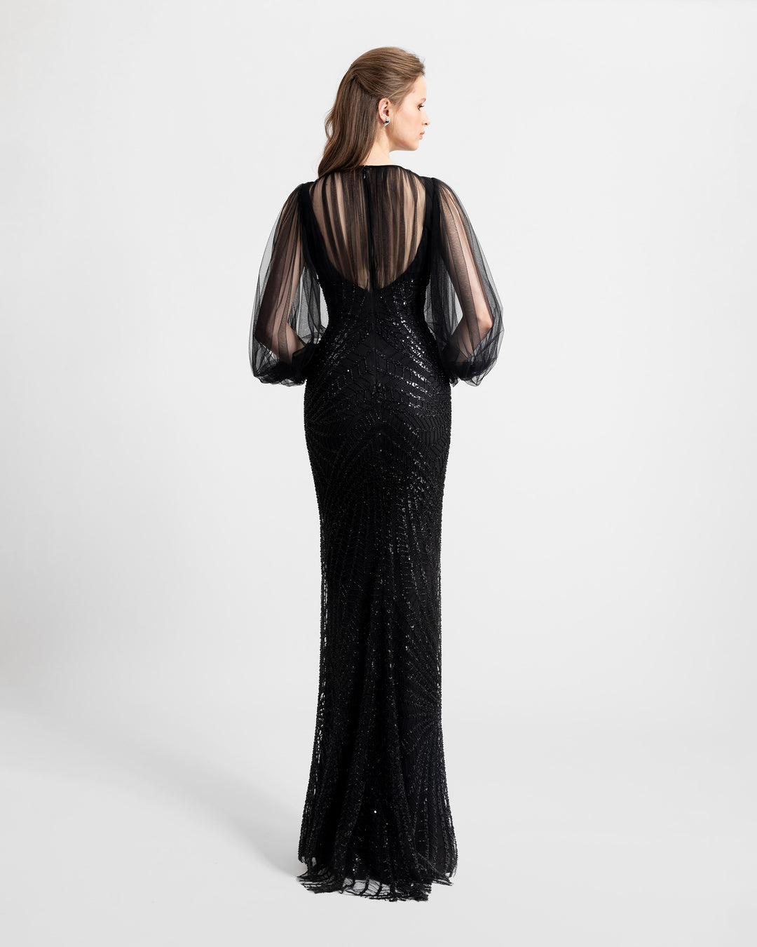 An open-back fitted black evening dress overlayed with a long sleeved draped tulle top and a fully embroidered see-through fabric from the the lower bust down.