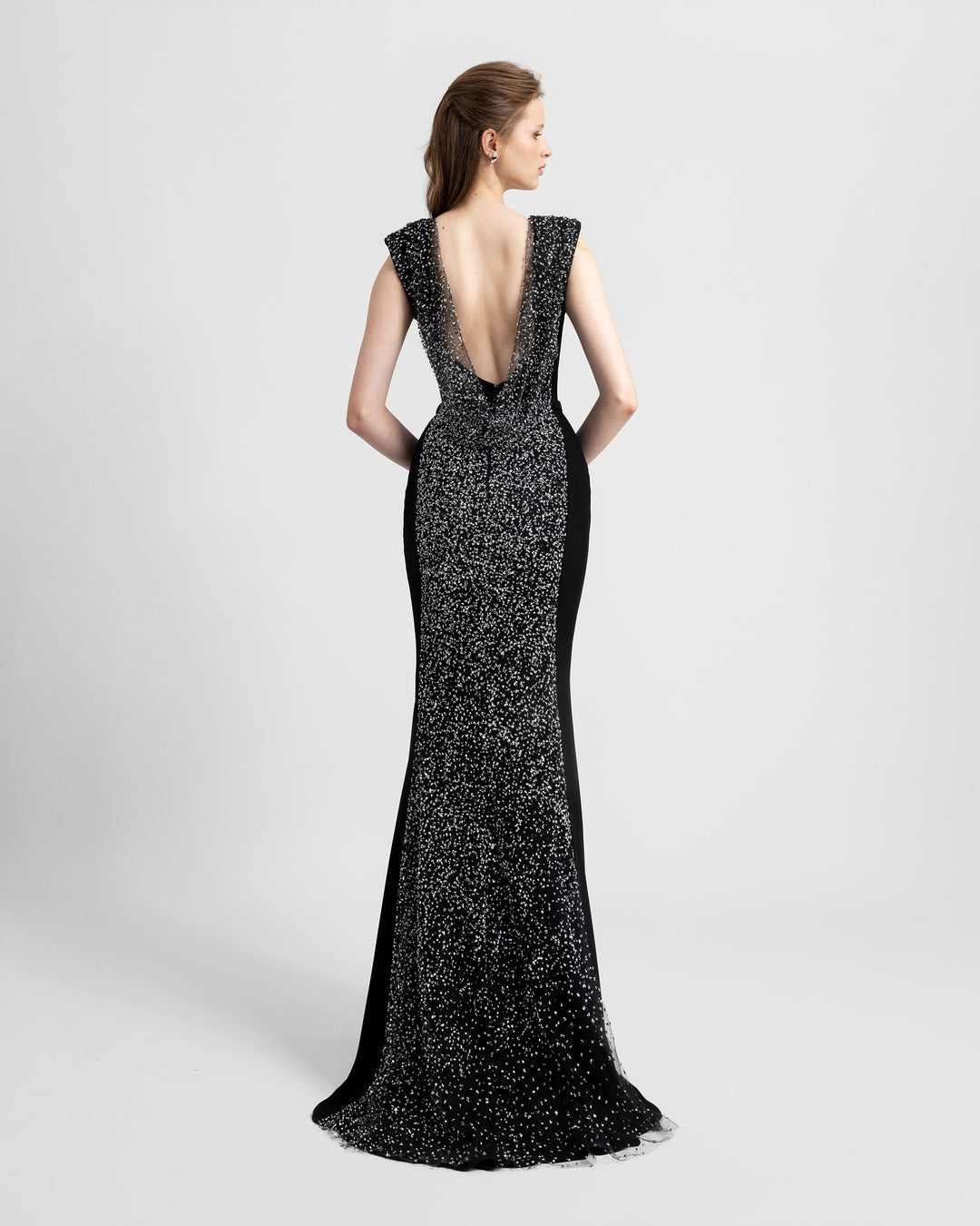 The back of a padded shoulders slim-cut black evening dress featuring an open draped back and a full back overlayed with beaded see-through fabric. 