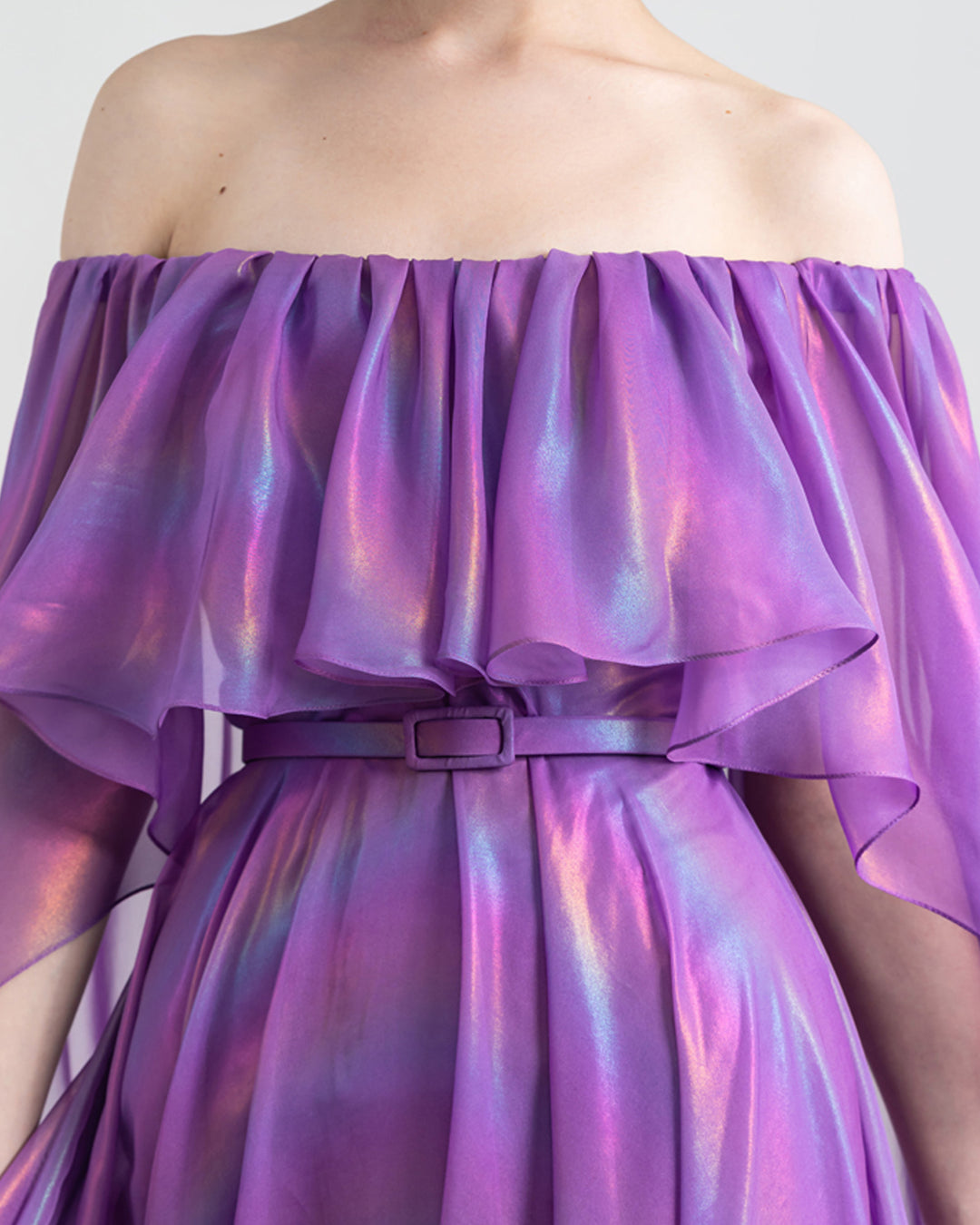 A close-up of an off-shoulders cape like sleeves, flared cut purple evening dress with ruffled details and a belt..