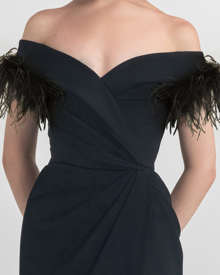 A close-up of a feathers off shoulder slim cut crepe long black dress with draping details on the waist and an open slit on the side.