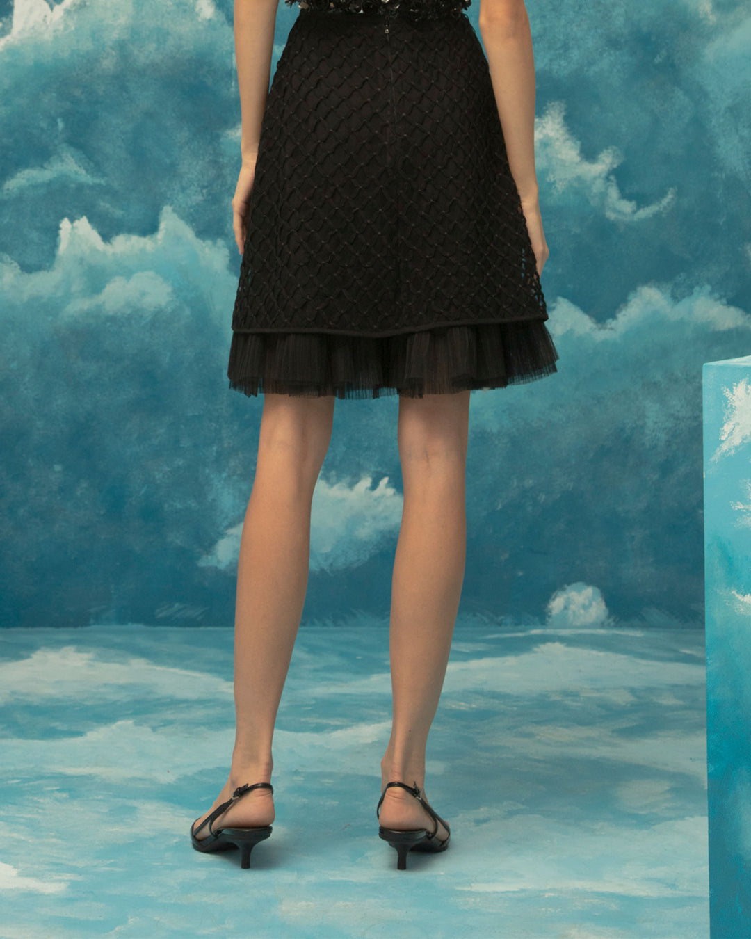 The back of a straight cut, embroidered fishnet short black skirt with ruffled details.