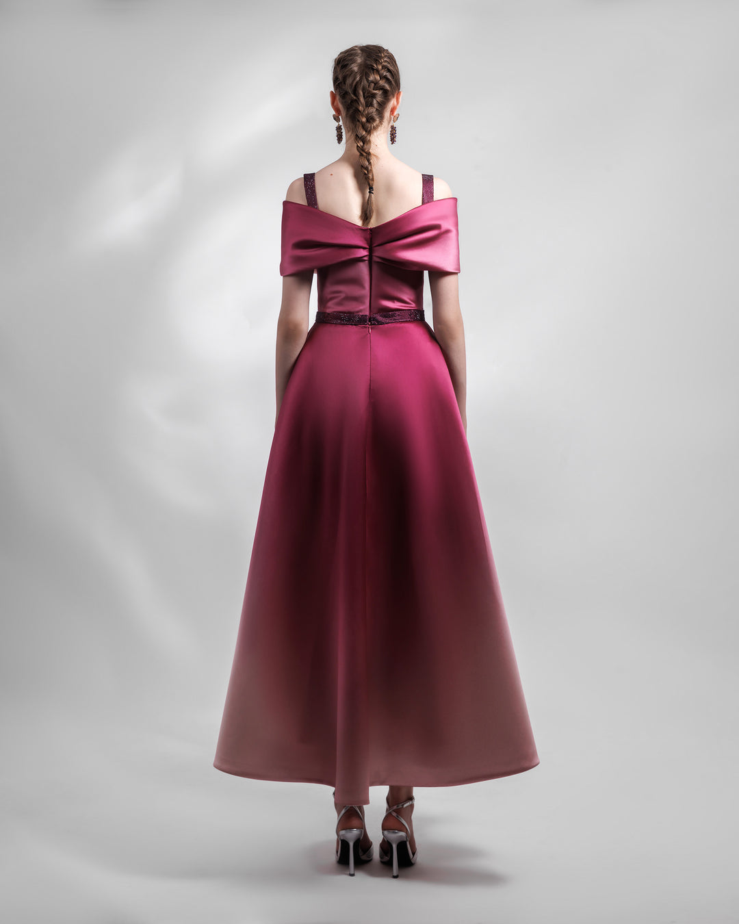 The back of a heavy satin midi dress in pink and burgundy hues, with a bow-like draped bodice, beaded straps and belt, and a pleated skirt.