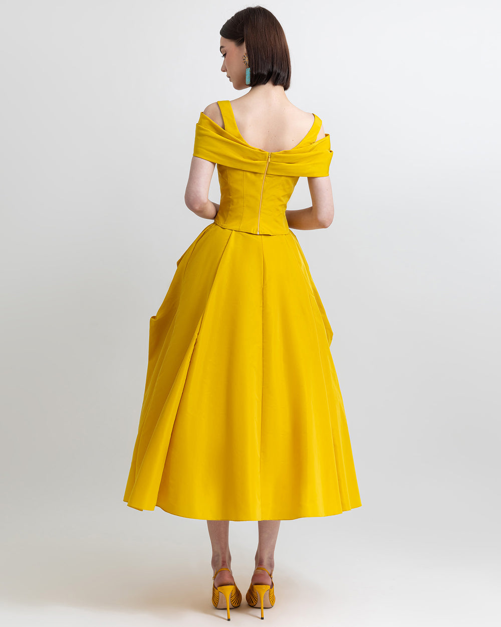 The back of an evening wear outfit featuring an asymmetrical bow-design taffeta corset top and a wide cut taffeta skirt with draping details, all in yellow color.