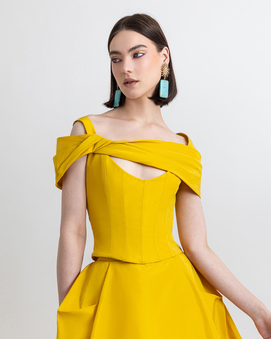 A close-up of an evening wear outfit featuring an asymmetrical bow-design taffeta corset top and a wide cut taffeta skirt with draping details, all in yellow color.