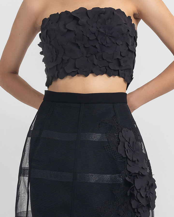 A close-up of an evening wear set featuring a strapless black cropped-top with laser-cut details, paired with a cage-like black skirt with flower details.