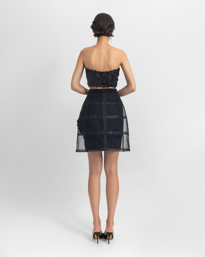 The back of an evening wear set featuring a strapless black cropped-top with laser-cut details, paired with a cage-like black skirt with flower details.