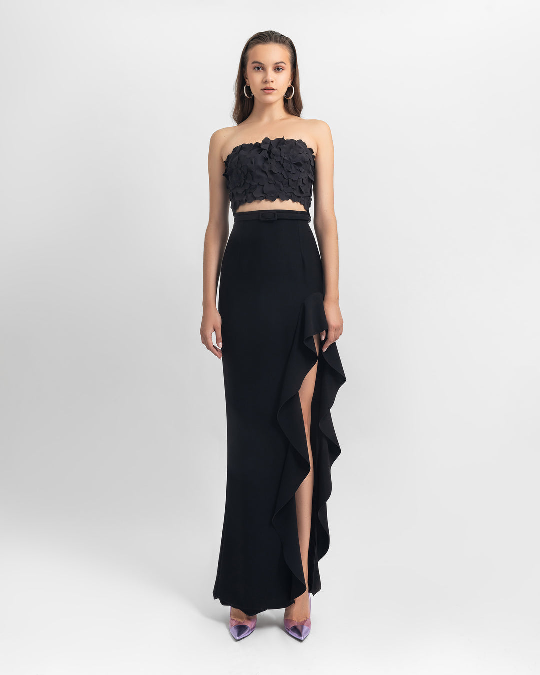 A black evening wear look featuring a strapless black cropped-top with laser cut details paired with an asymmetrical crepe skirt with ruffled details and a slit on the side.