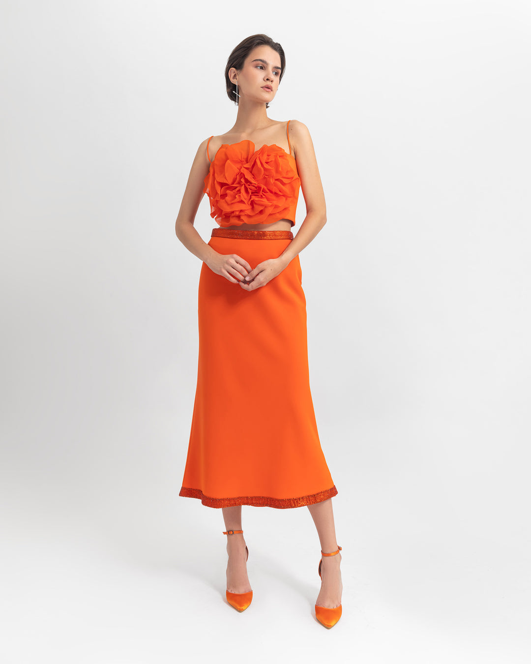 An evening wear orange set featuring a flower draped chiffon cropped top paired with a straight cut crepe orange skirt with sequin details on the waist and hemline.