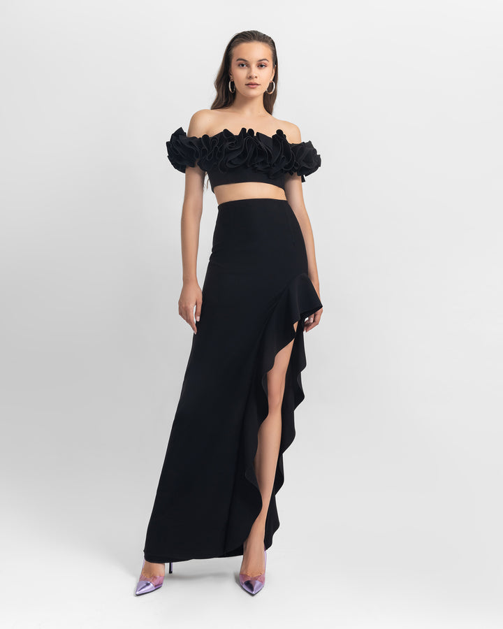 A black evening wear look featuring an off-shoulders ruffled crepe crop top paired with an asymmetrical crepe skirt with ruffled details and a slit on the side.