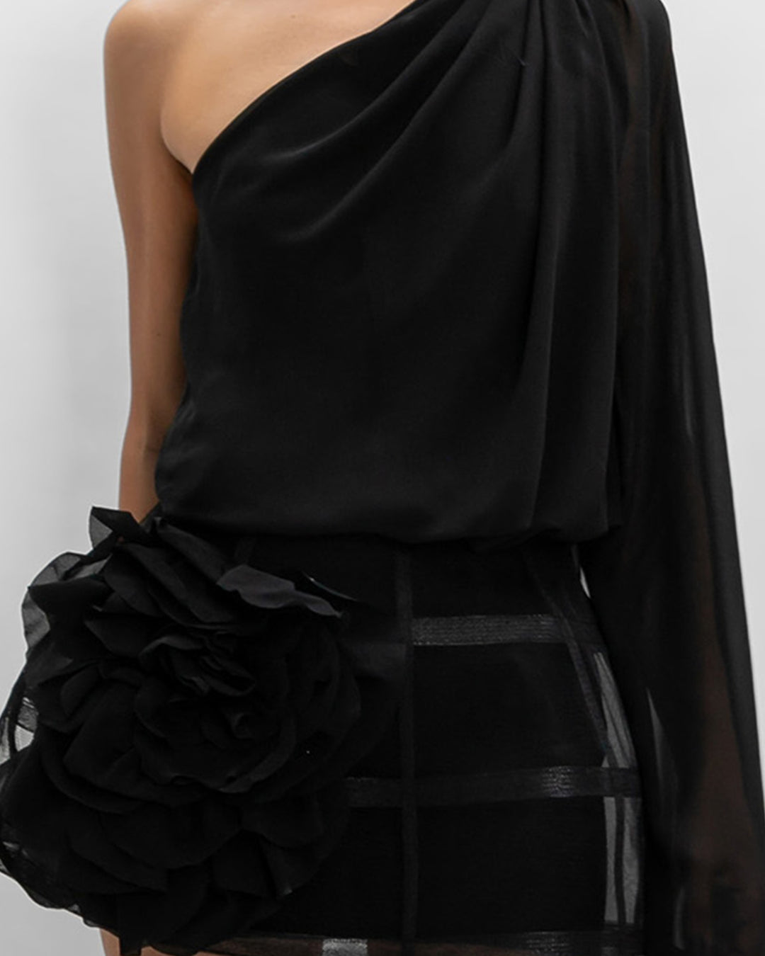 A close-up of a black evening wear set featuring an asymmetrical loose-cut chiffon top, paired with a short cage-like skirt with flower details on the side.