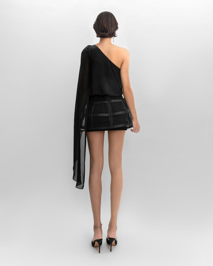 The back of a black evening wear set featuring an asymmetrical loose-cut chiffon top, paired with a short cage-like skirt with flower details on the side.