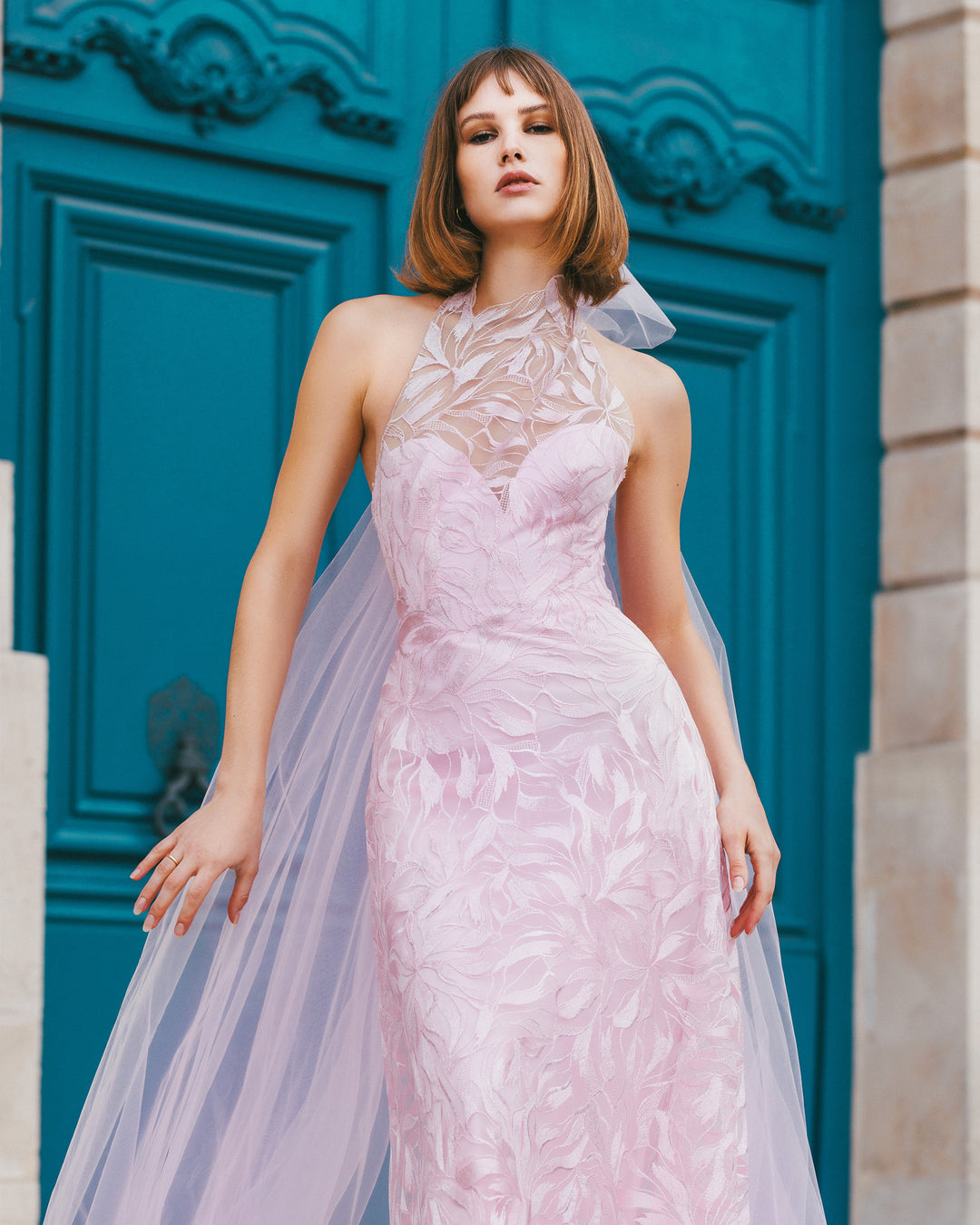 A flared cut embroidered fabric long pink dress with an overlayed halter neckline and a backless design, paired with a detachable tulle cape.